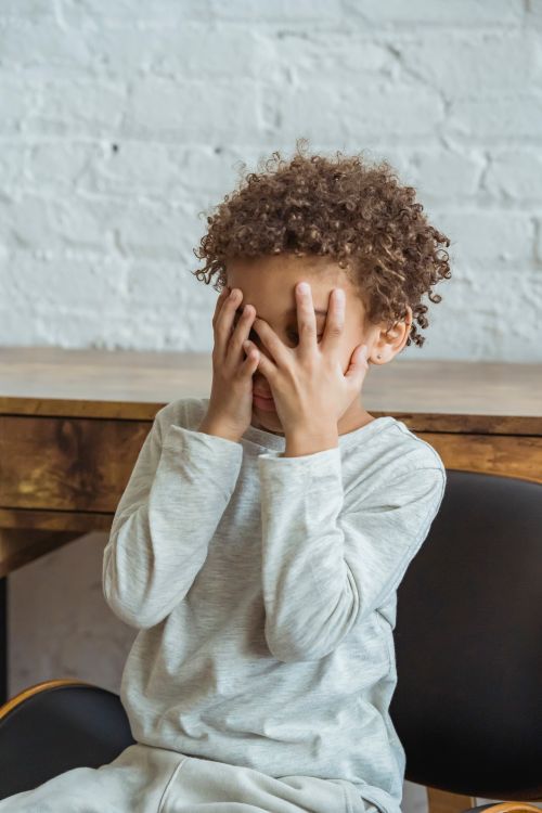 Free Anonymous frustrated African American boy with curly hair in casual clothes covering face with hands while sitting in light room Stock Photo