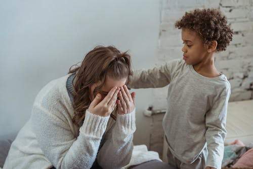 Free Caring African American son touching shoulder of upset faceless mother covering face while sitting in light room near wall at home Stock Photo