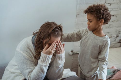 Caring African American son touching shoulder of upset faceless mother covering face while sitting in light room near wall at home