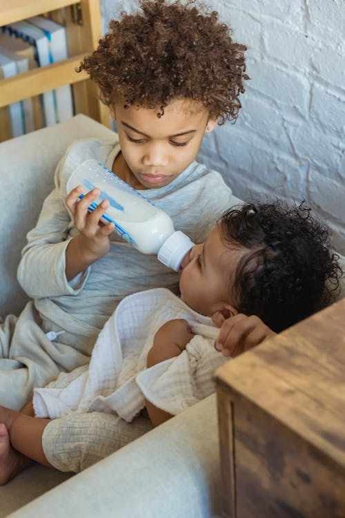 From above of caring African American baby bottle feeding adorable black toddler while sitting in armchair near wall at home