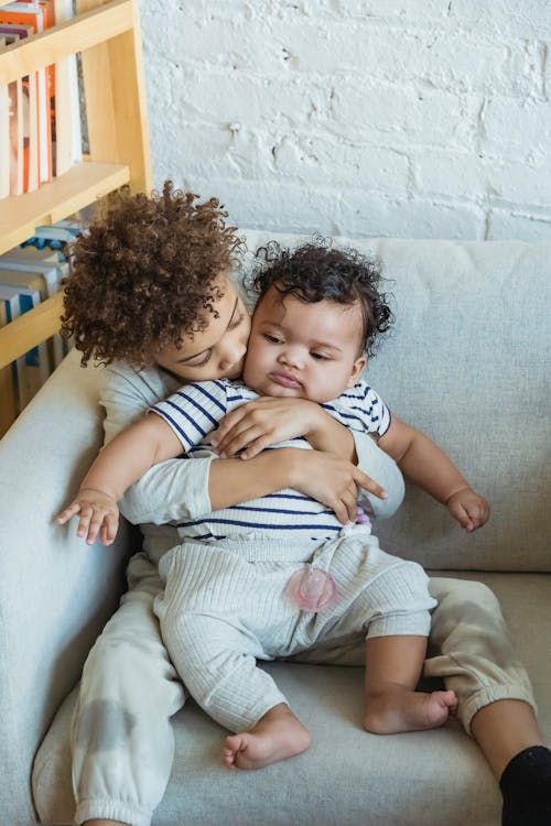 Free Tender African American boy cuddling adorable little baby while sitting on comfortable sofa near wall in light room at home Stock Photo