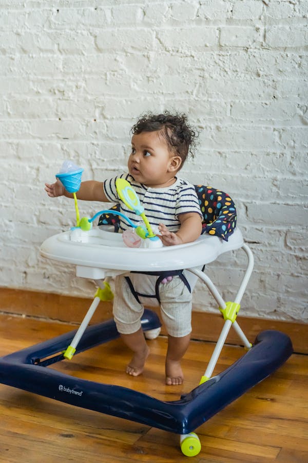 Full body of barefoot African American toddler looking away while sitting in baby walker with toys near white wall at home