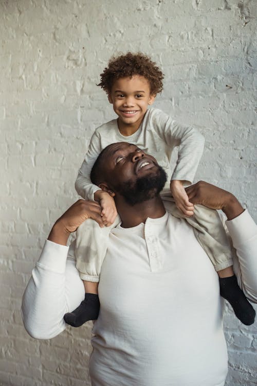 Cheerful African American father looking at glad black son sitting on shoulders while having fun near wall in light room