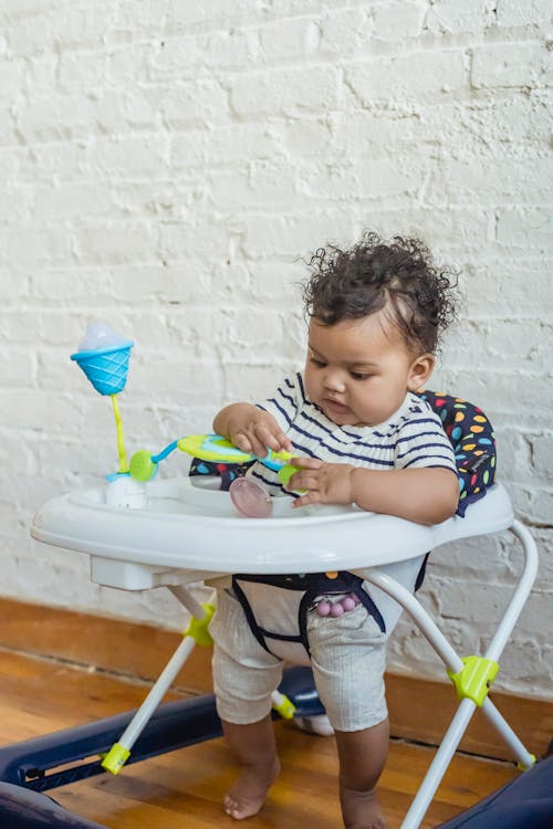 Free Curious little barefoot African American baby sitting in walker and playing with toy near white wall in room at home Stock Photo