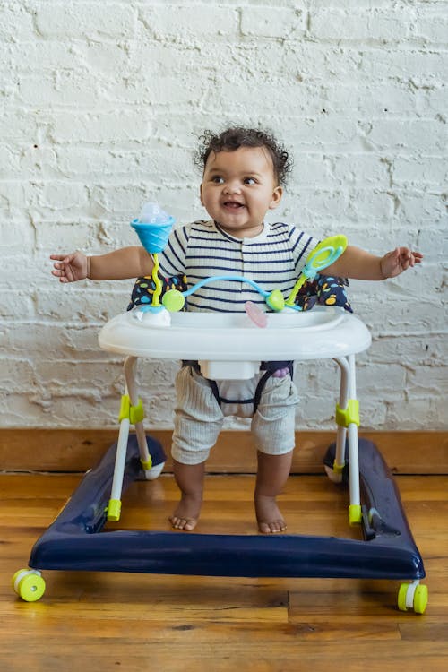 Free Full body of adorable barefoot African American child looking away while sitting in baby walker near wall at home Stock Photo