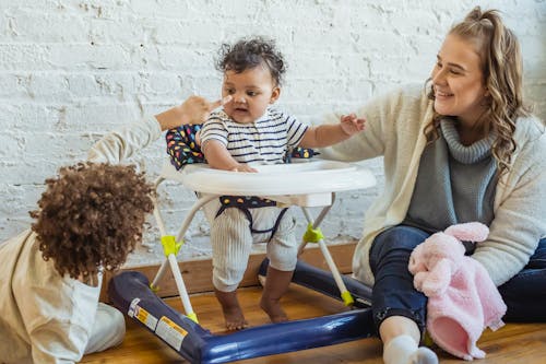Free Happy mother with toy playing with black toddler in baby walker and boy while sitting on floor near wall at home Stock Photo