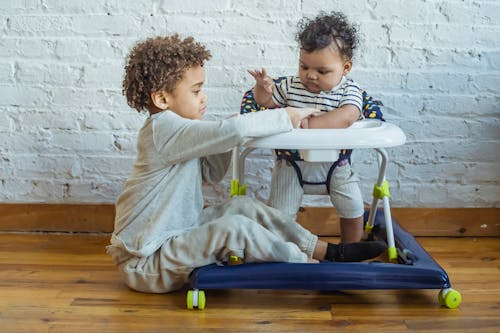 Full body of content African American boy sitting near small black toddler in baby walker while playing on floor near wall