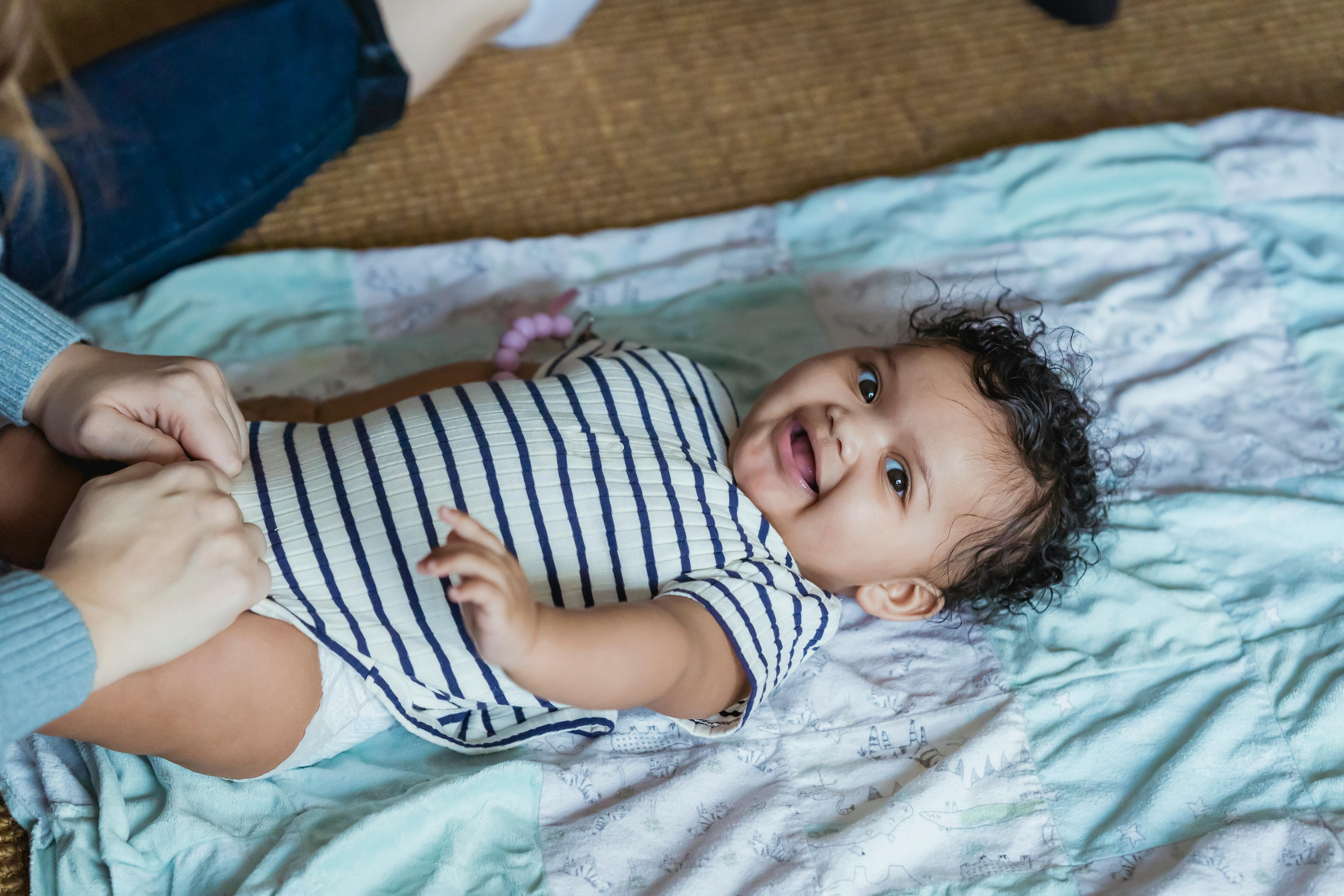 A happy baby in a striped onesie being dressed on a blanket indoors.