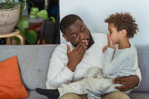 African American man with child in casual outfit covering mouth with hand on couch with pillows while looking at each other in light apartment