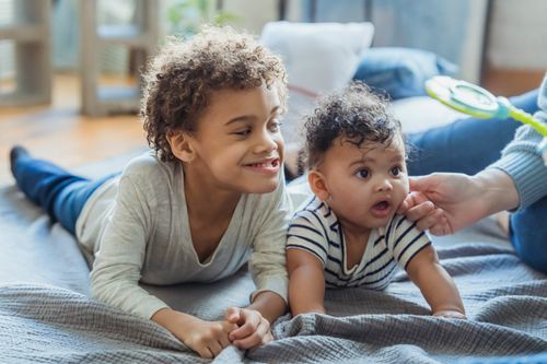 Free Multiracial children in casual clothes grimacing while lying on textile on floor while mother pulling hand to baby cheek in light room Stock Photo