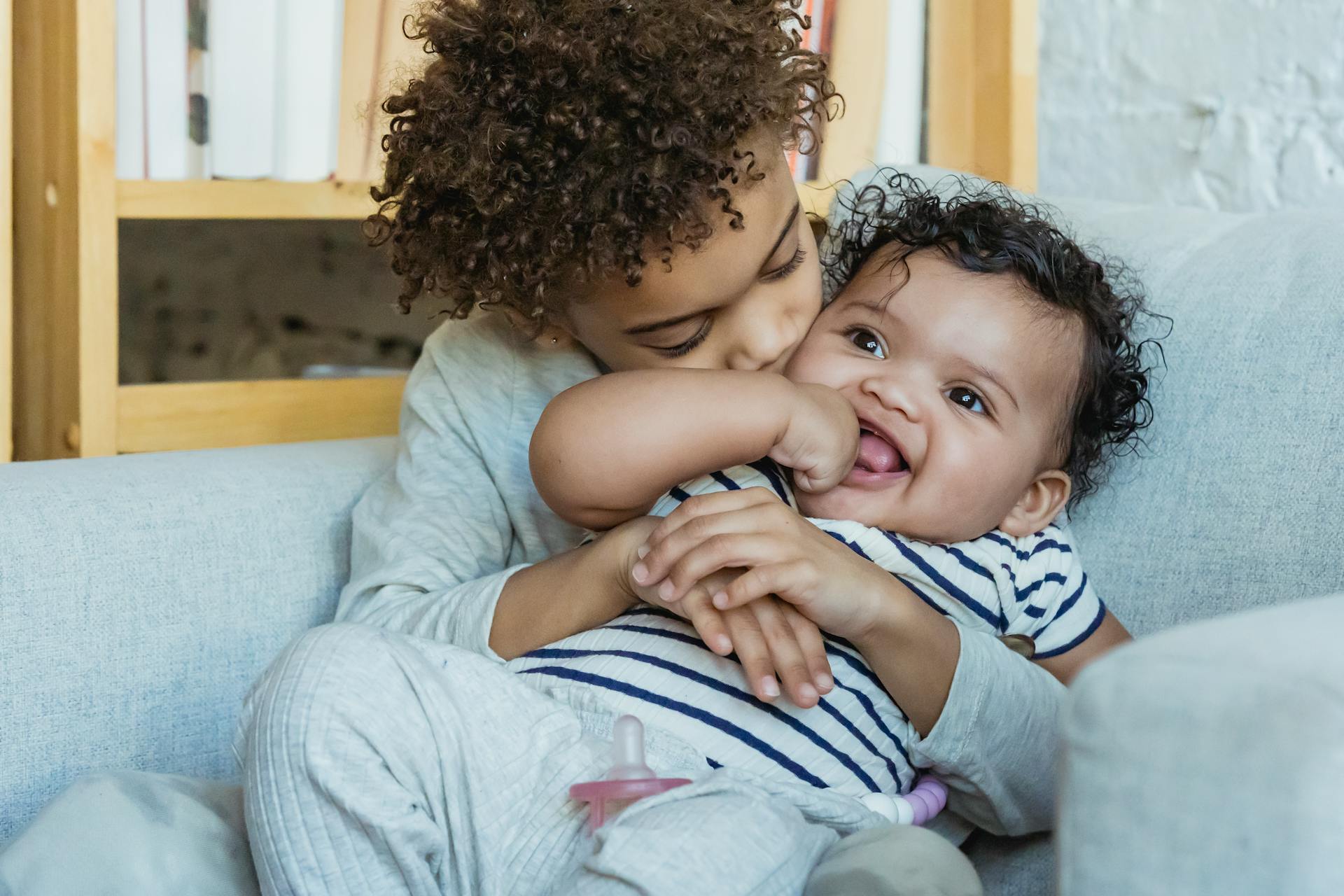 African American boy with curly hair embracing and kissing adorable smiling little baby while sitting in comfortable armchair