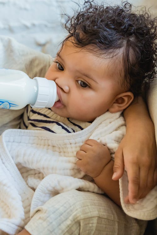 Adorable African American baby with curly hair drinking milk from bottle while sitting on sofa wrapped in blanket on blurred textured background and looking away