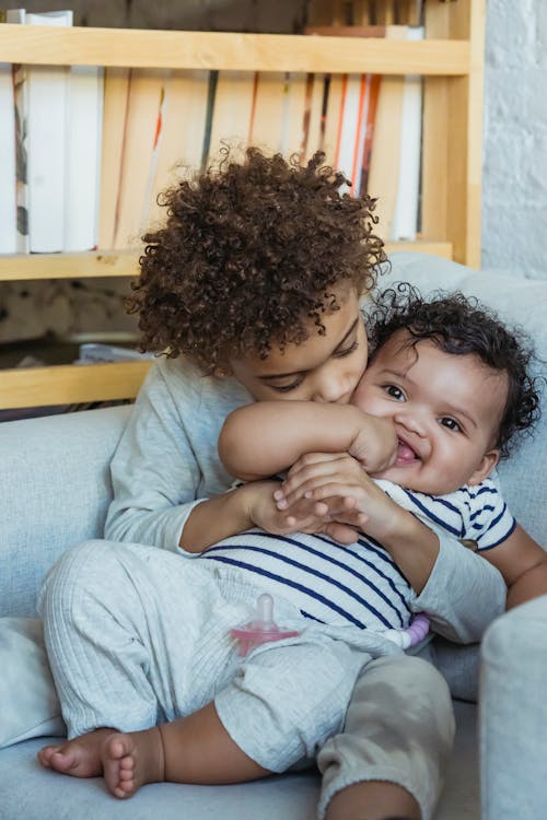 African American child with curly hair hugging and kissing little smiling baby while sitting on comfortable sofa at home