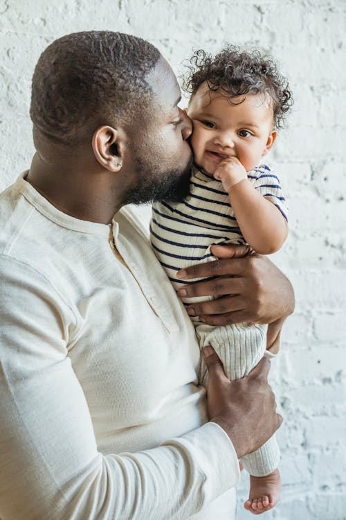 Caring adult African American father in white casual shirt holding and kissing adorable baby cheek against white wall