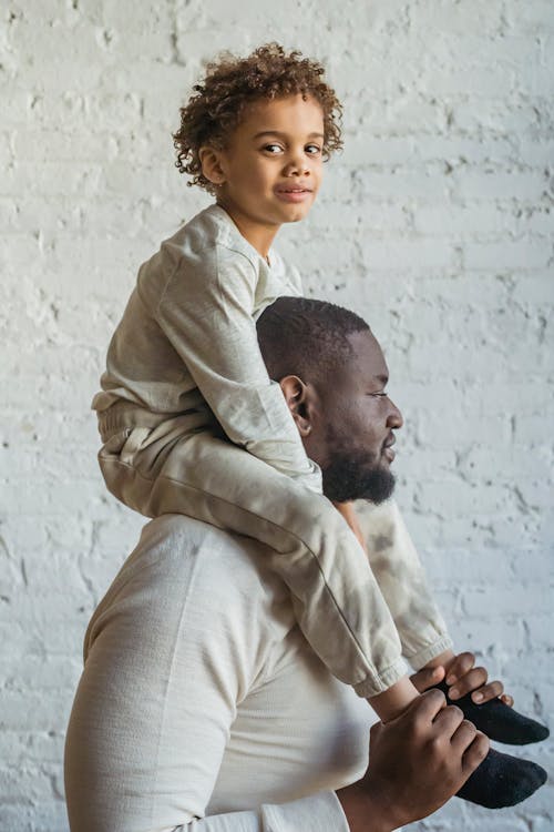 Side view focused African American male in casual wear carrying content son on shoulders against white wall in light room
