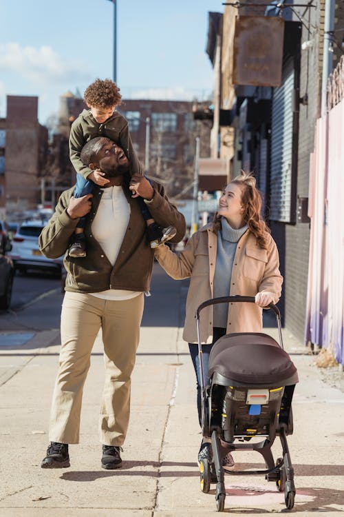 Full body cheerful black father carrying son on shoulders and walking on city street near happy wife with stroller