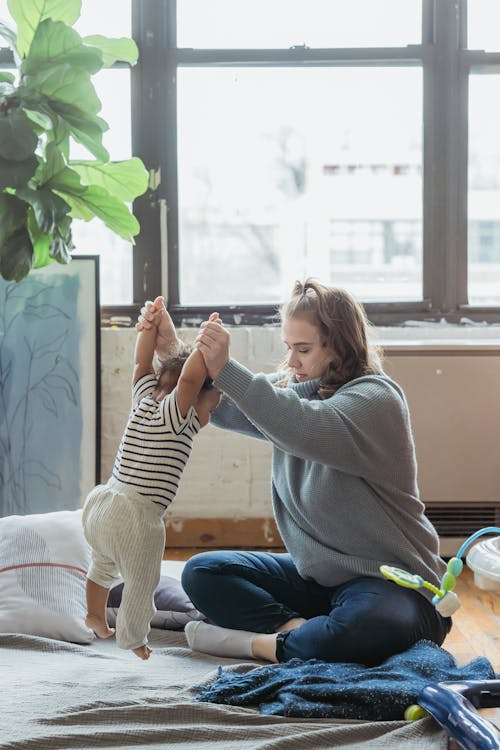 Full body positive mother in casual clothes teaching adorable toddler to walk while sitting on floor with legs crossed in light room