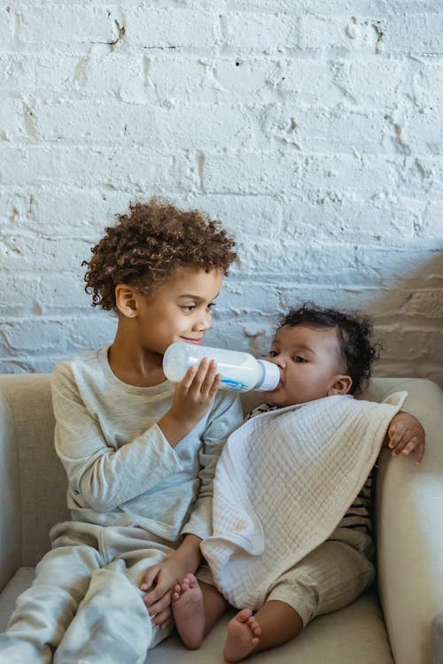 Content ethnic boy in domestic wear feeding adorable sibling with milk bottle while sitting together on comfy armchair