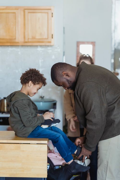Free Cheerful African American boy smiling while father helping with boots at home in daytime Stock Photo