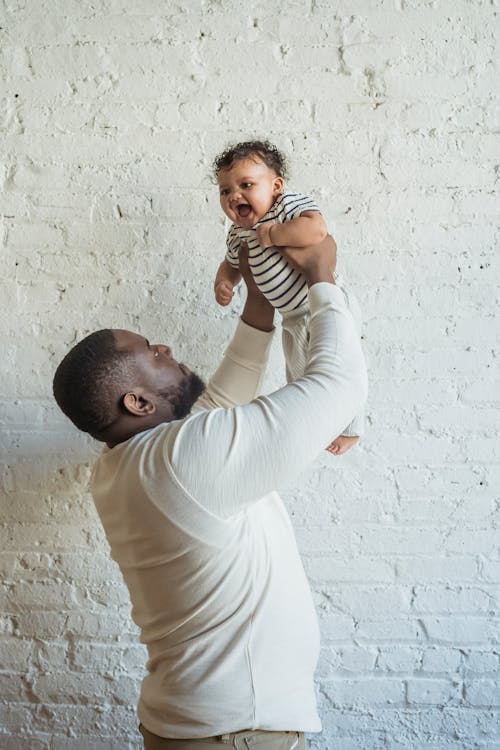 Side view of African American father with adorable small toddler in arms near white brick wall