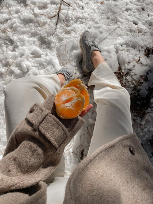 Free From above of anonymous female in warm coat and ripe peeled tangerine in hand sitting in winter nature with snowy ground Stock Photo