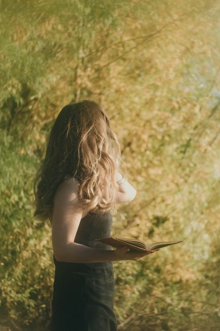 A woman standing outdoors holding a book, surrounded by lush greenery on a sunny day.