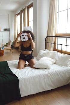 Woman sitting on bed, holding facial mask, ready for skincare in bright room.