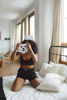 Relaxed woman holding a face mask while kneeling on bed in cozy bedroom.