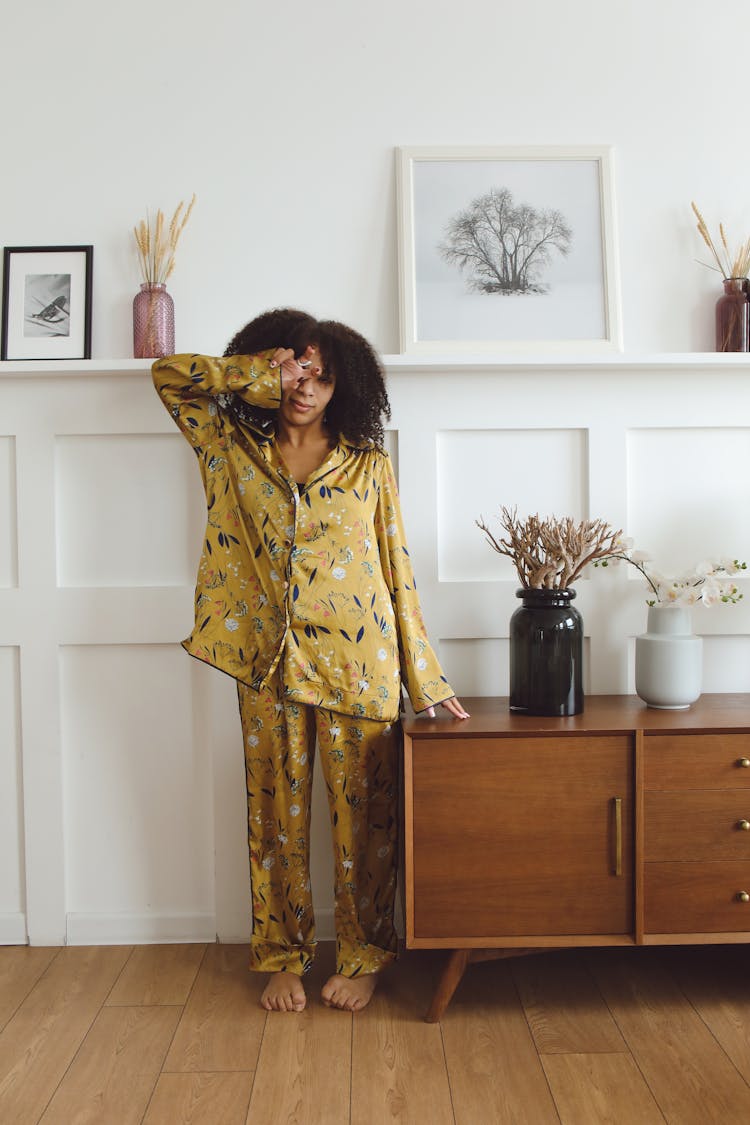 Woman In Pajamas Standing Beside The Wooden Cabinet With Flower Vase