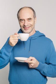Elderly man in blue hoodie drinking coffee and smiling indoors.