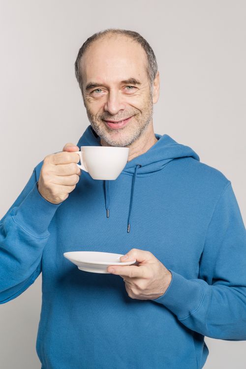 Free Elderly man in blue hoodie drinking coffee and smiling indoors. Stock Photo