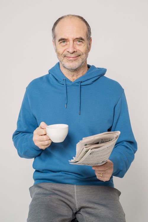 Free Senior adult in blue hoodie reading newspaper while holding a coffee cup indoors. Stock Photo