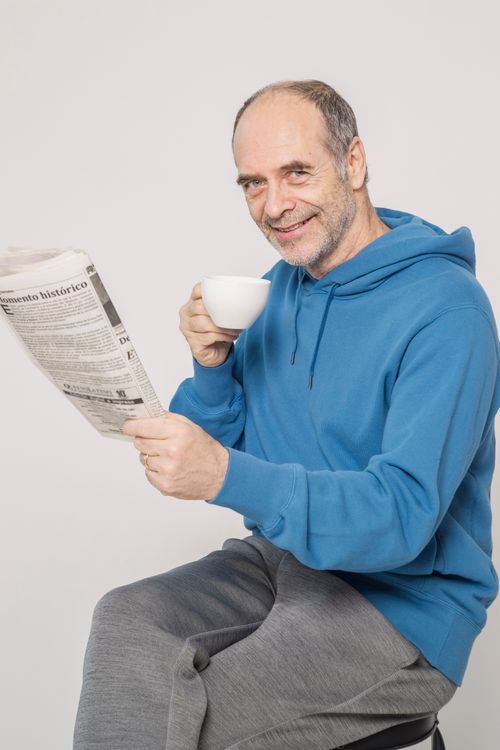 Free Smiling senior adult in a blue hoodie reading a newspaper while drinking coffee indoors. Stock Photo