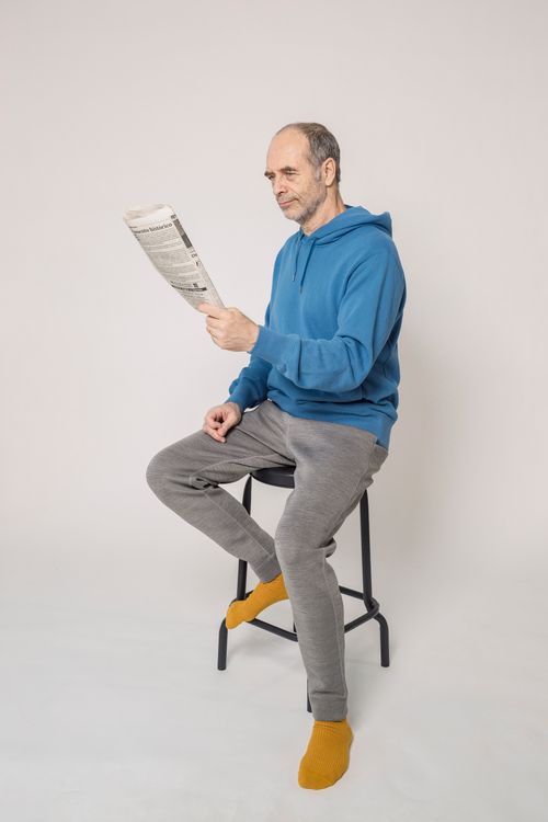 Free Elderly man in casual hoodie reading a newspaper indoors, seated on a stool. Stock Photo