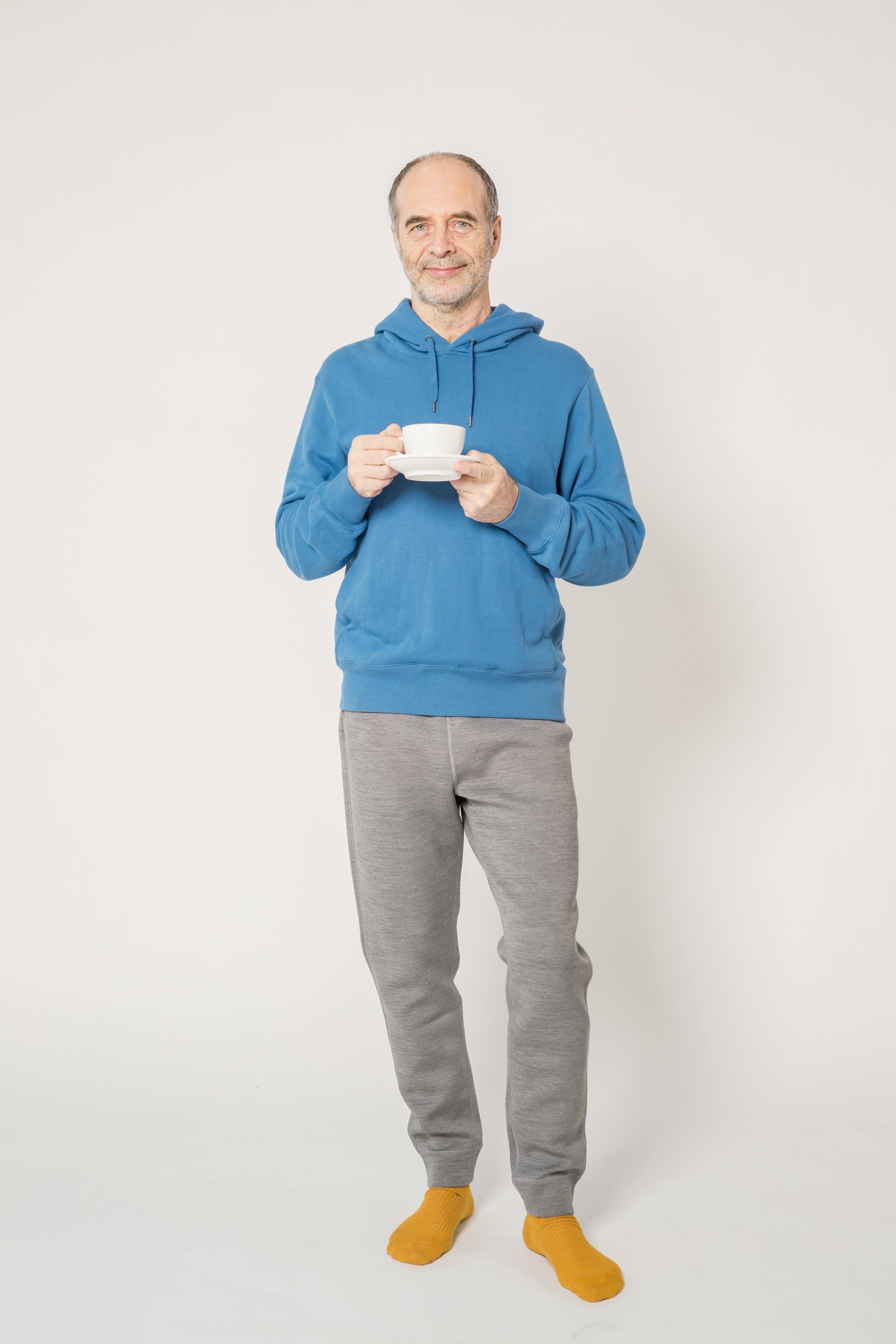 Senior man wearing a blue hoodie, holding a cup indoors, smiling casually.