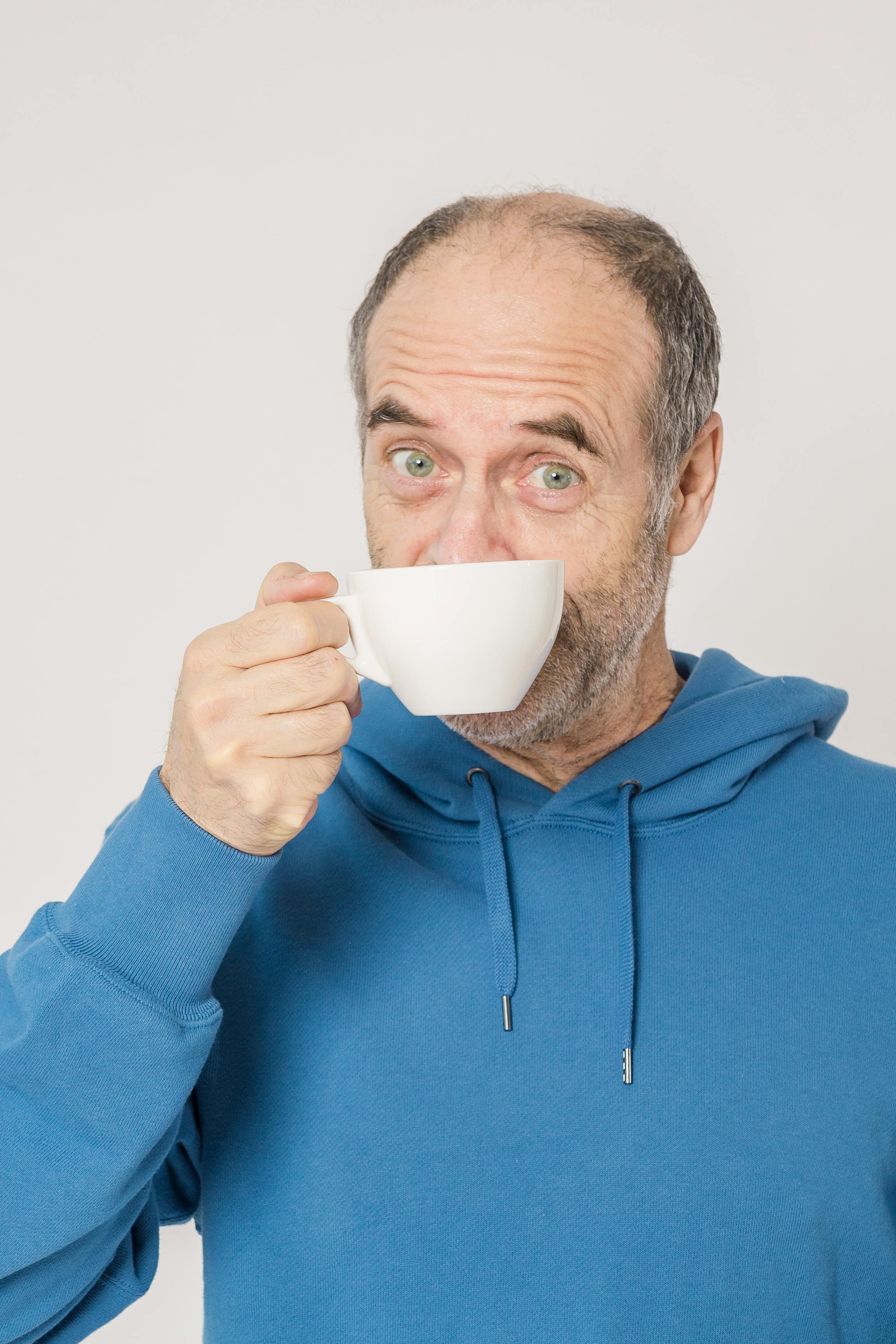Elderly man in blue hoodie enjoying a hot drink indoors. Relaxed and casual setting.