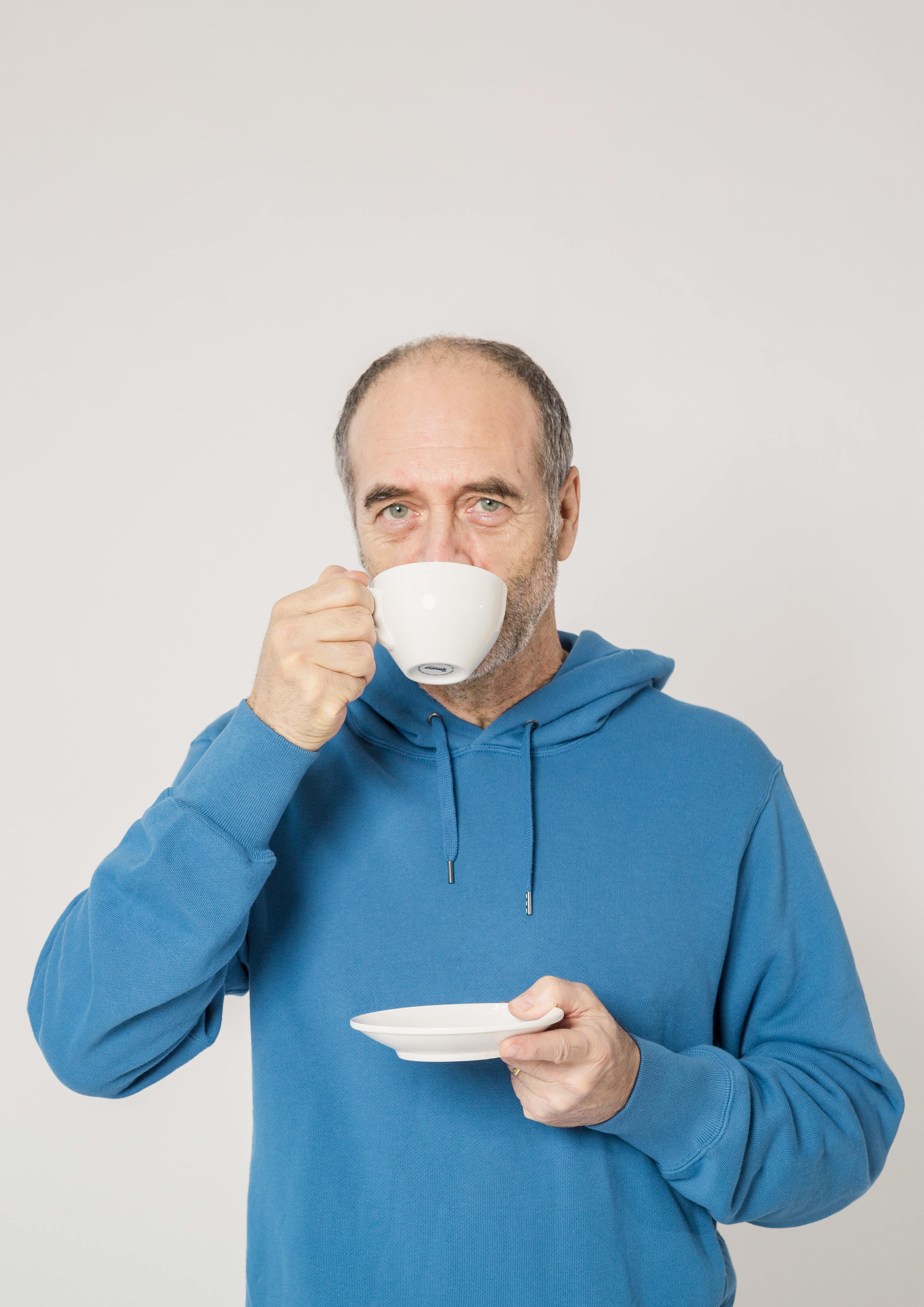 Elderly man in a blue hoodie drinking from a cup, enjoying a moment of relaxation.