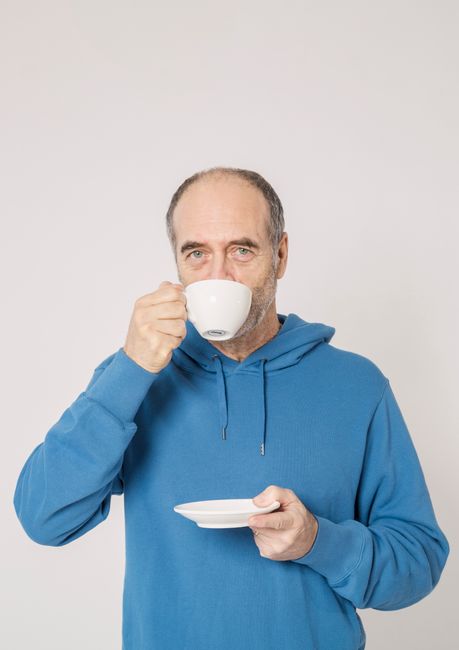Elderly man in a blue hoodie drinking from a cup, enjoying a moment of relaxation.