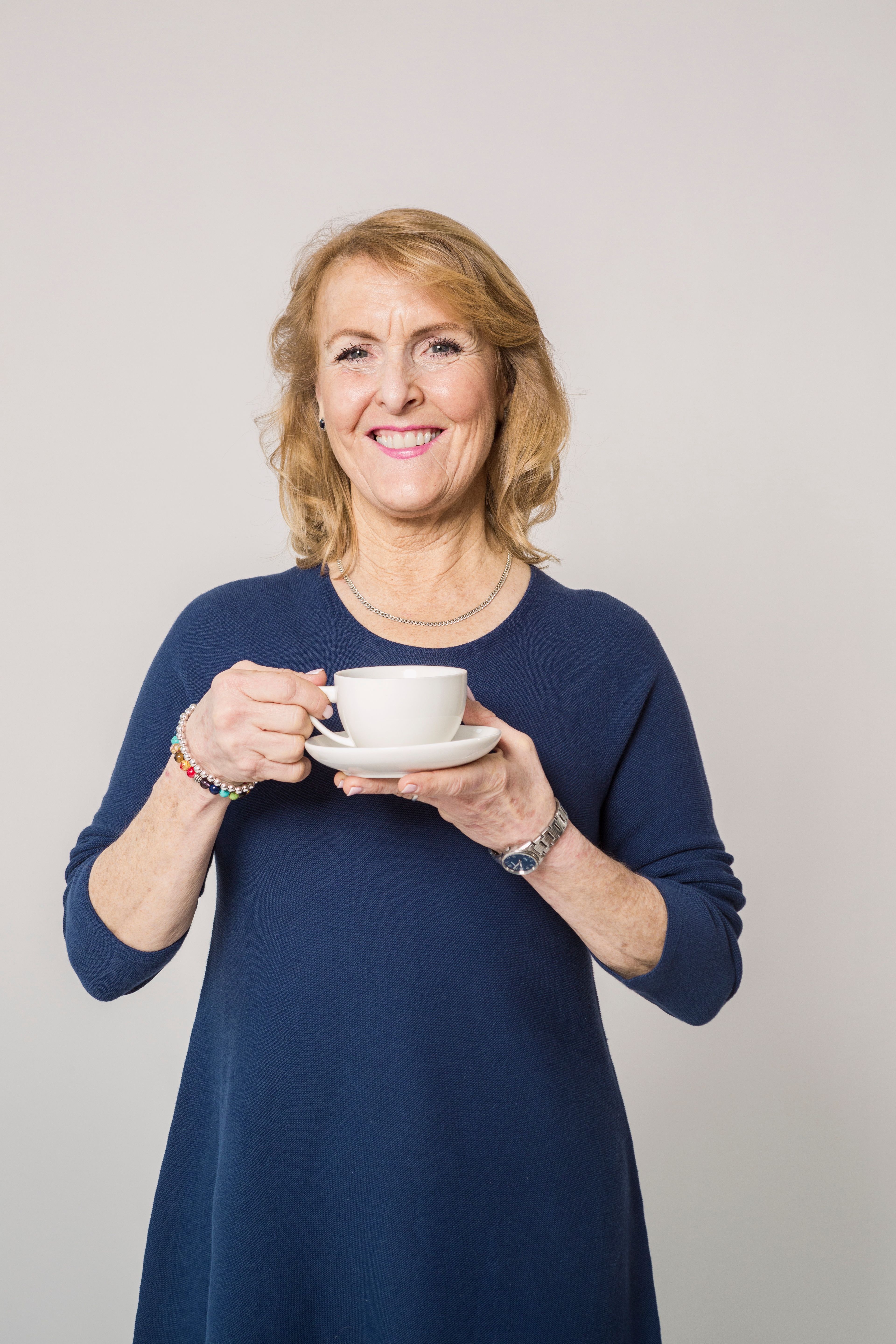 A cheerful senior woman in a blue dress enjoying a cup of coffee indoors, exuding warmth and relaxation.