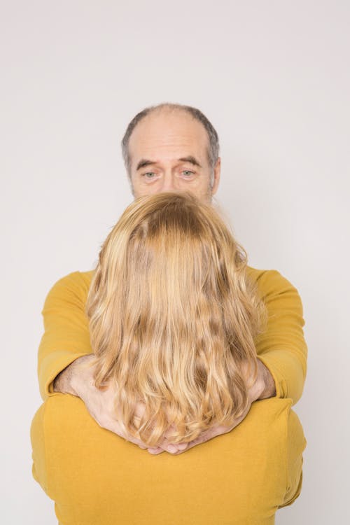 Back view of unrecognizable woman with elderly husband in similar outfits embracing and looking at each other against white background
