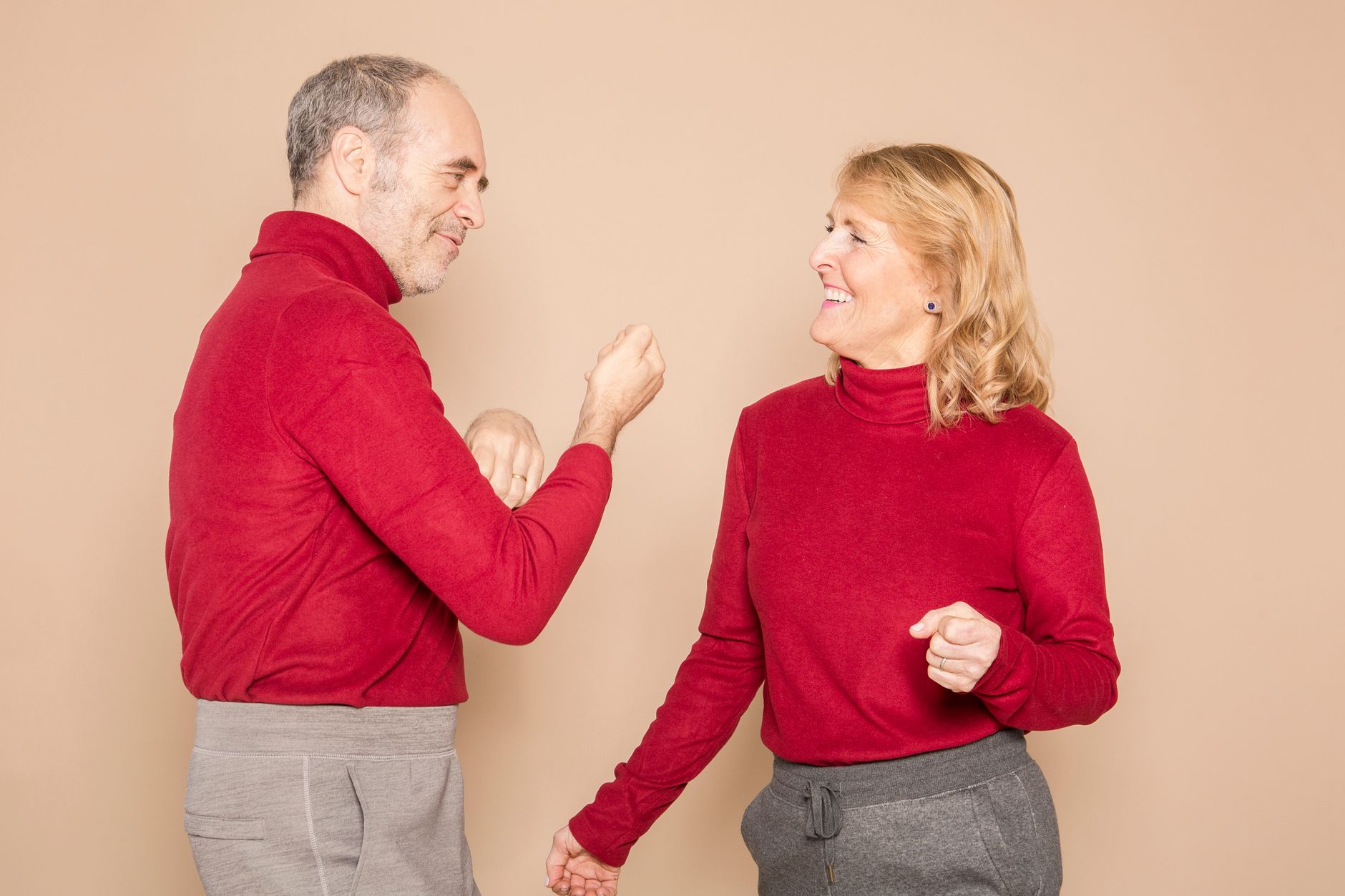 Joyful elderly couple dancing together in matching red sweaters against a neutral backdrop.
