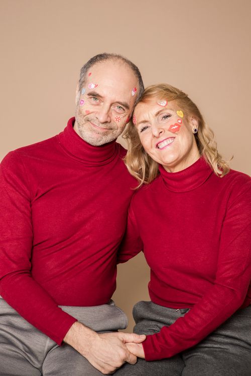 Free Smiling senior couple in red sweaters with heart stickers on their faces, sitting together indoors. Stock Photo