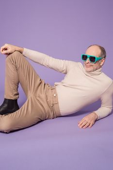 Elderly man wearing sunglasses and a sweater poses confidently against a purple background.
