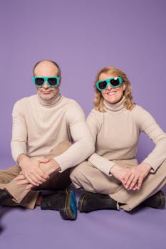Elderly couple in beige outfits with teal sunglasses, smiling on a purple backdrop.