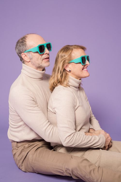 Free Charming senior couple in matching beige sweaters and sunglasses, embracing joyfully against a purple studio backdrop. Stock Photo