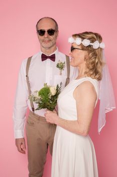 Charming senior couple in wedding attire posing joyfully against a pink background.