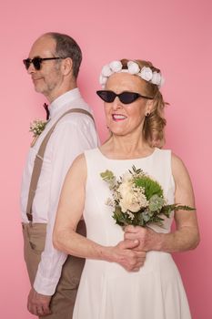 Elderly couple in elegant wedding attire posing playfully in a studio with a pink background.