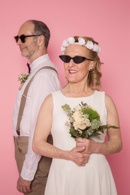 Free Elderly couple in elegant wedding attire posing playfully in a studio with a pink background. Stock Photo
