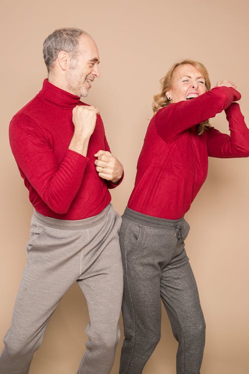 Joyful mature couple in similar red sweater dancing happily together while having fun in beige studio