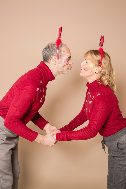 Free Side view of positive elderly couple in similar clothes and heart party headbands holding hands and pouting lips while standing in studio during Saint Valentines Day Stock Photo